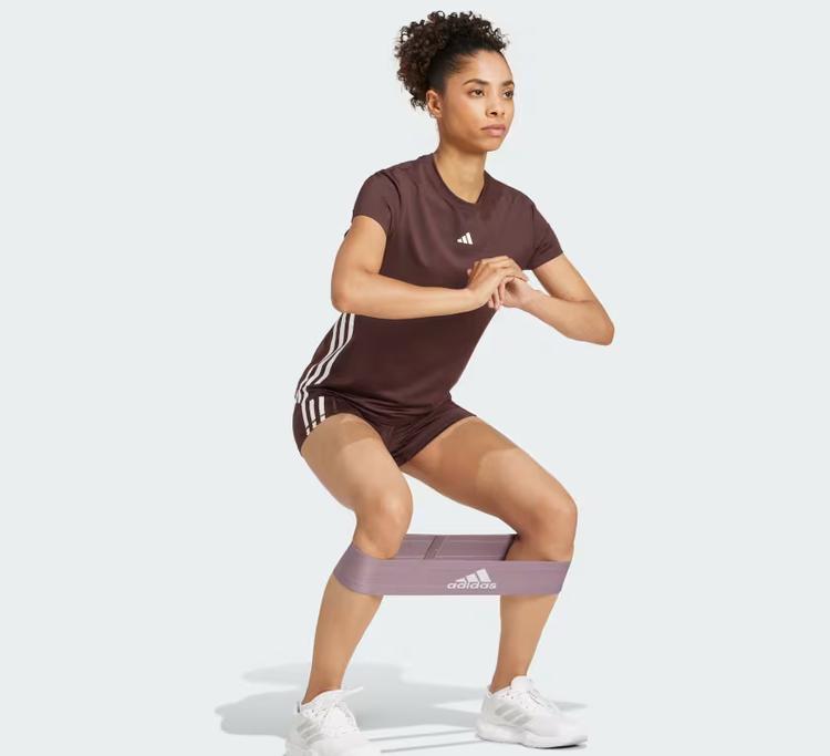 Een vrouw draagt een bruin sportshirt en bruine sportshort van Adidas en voert een squat uit met een weerstandsband